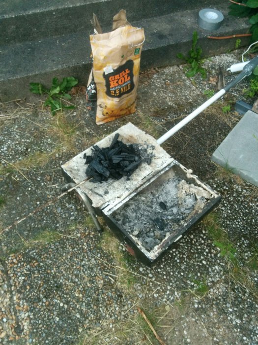 En enkel murad utegrill med grillkol och en tom grillkolsäck intill på en grusig mark.
