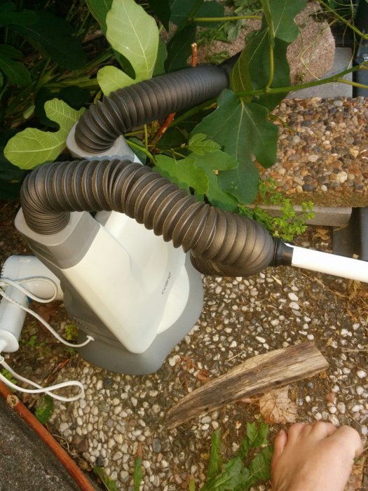 Flexibla ventilationsrör anslutna till en vit luftavfuktare utomhus omgiven av grus och växtlighet.