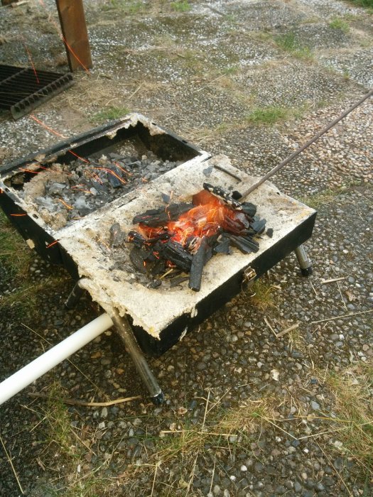 En utomhusgrill med brinnande träkol och en järnspett över glöden, placerad på en grusig mark.