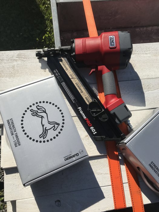Nytt köpt verktyg, en röd spikpistol, ovanpå kartonger med tillbehör, placerade på vit träyta i solljus.