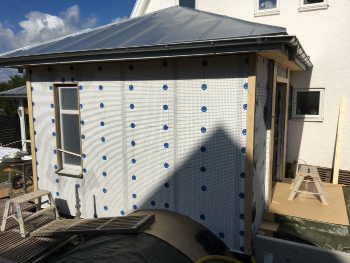 Husunderarbete med isoleringsmaterial och en fasad täckt av vindskyddsskiva och blå pluggar.