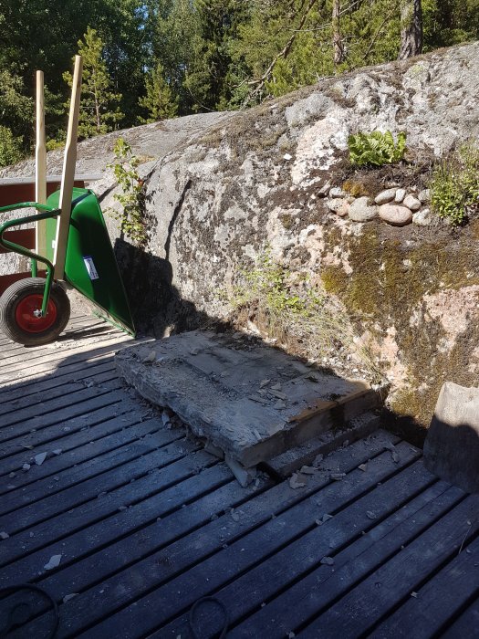 Resterna av en gammal grill på en träaltan med en grön skottkärra i bakgrunden.