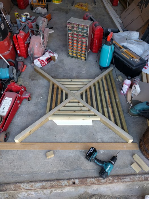 Pågående bygge av en stor trägrind på ett rörigt garagegolv med verktyg och byggmaterial omkring.