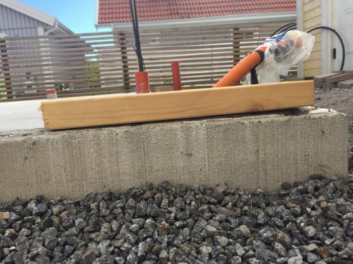 Ojämn betonggrund som sticker ut ovan kantelement bredvid en trälist och grus, med byggmaterial i bakgrunden.