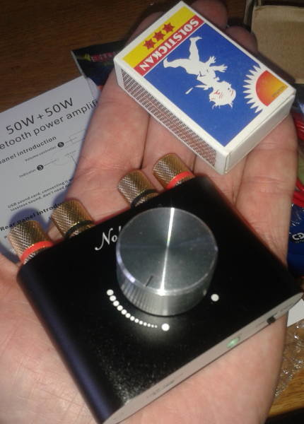 En hand håller en liten svart ljudförstärkare jämfört i storlek med en tändsticksask, med synliga kontakter och volymknapp.