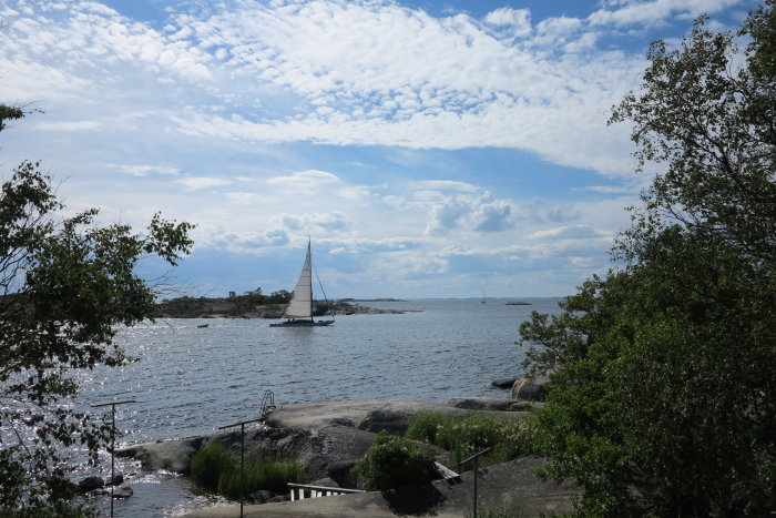 Segelbåt på glittrande vatten vid skärgård under molnig himmel omgivet av grönska.