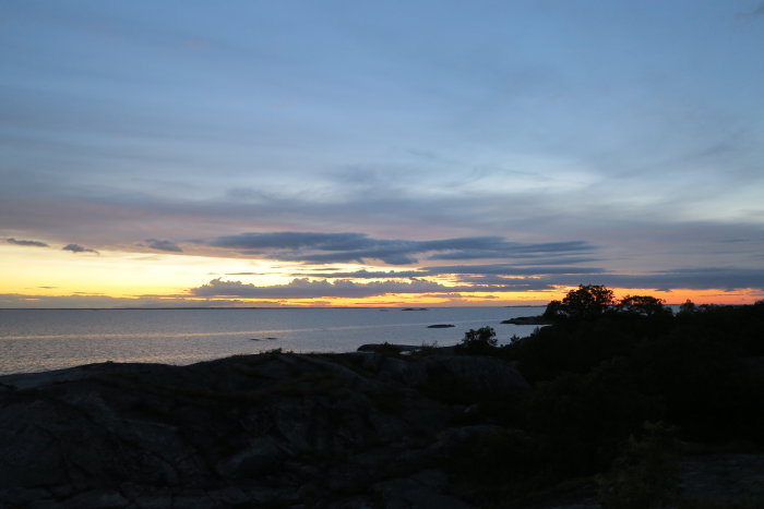 Solnedgång med färgglad himmel över hav och silhuett av klippor och träd.