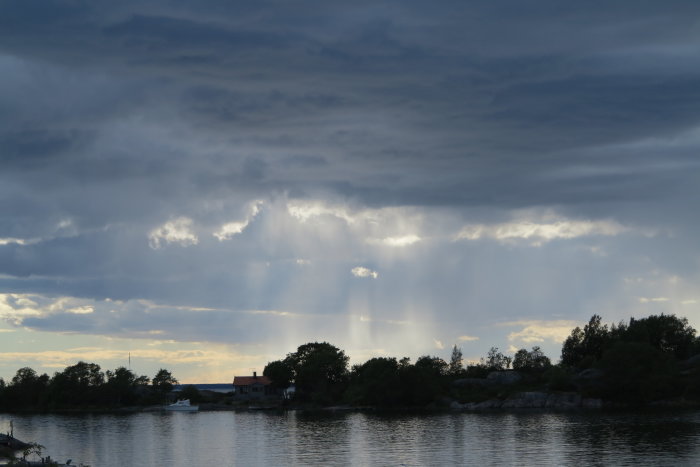 Molnigt landskap med solstrålar som bryter igenom molnen över en sjö med hus och träd.