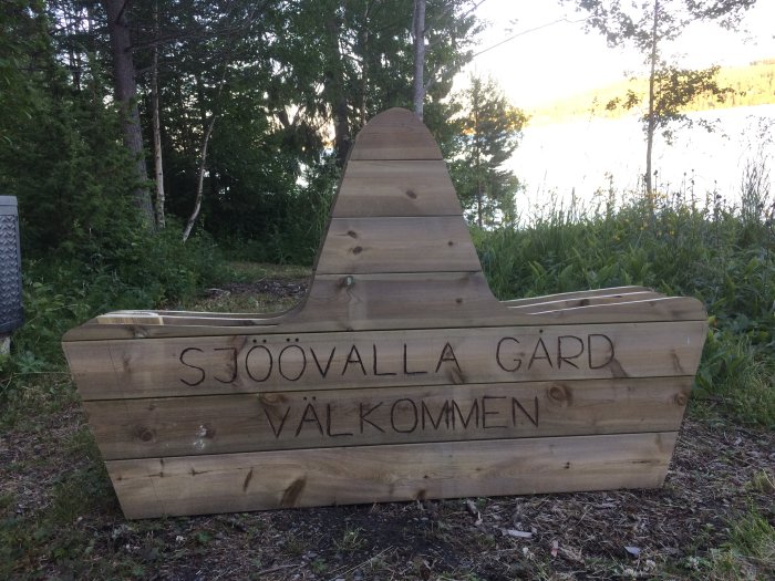 Påbörjad träsoffa med inskriptionen "SJÖÖVALLA GÅRD VÄLKOMMEN" framför en sjö med träd i bakgrunden.