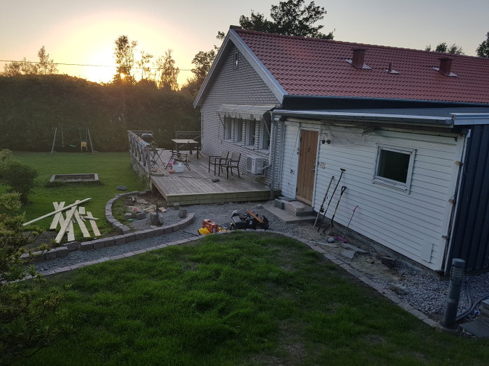 Solnedgång över en trädgård med nylagd stentrappa och verktyg, nära ett hus med terrass under renovering.