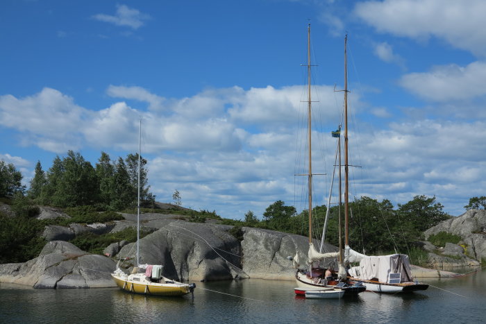 Två klassiska segelbåtar förtöjda vid klippor under en himmel med lätta moln.