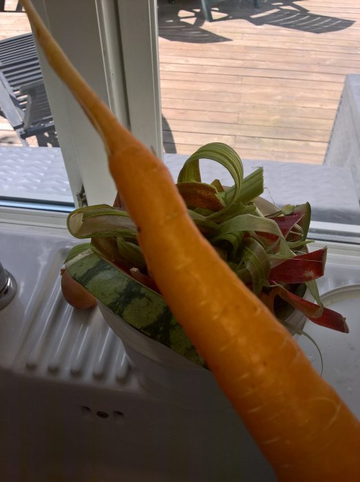 Lång morot nära kameran med komposthög av grönsaksrester i bakgrunden på en vit diskbänk.