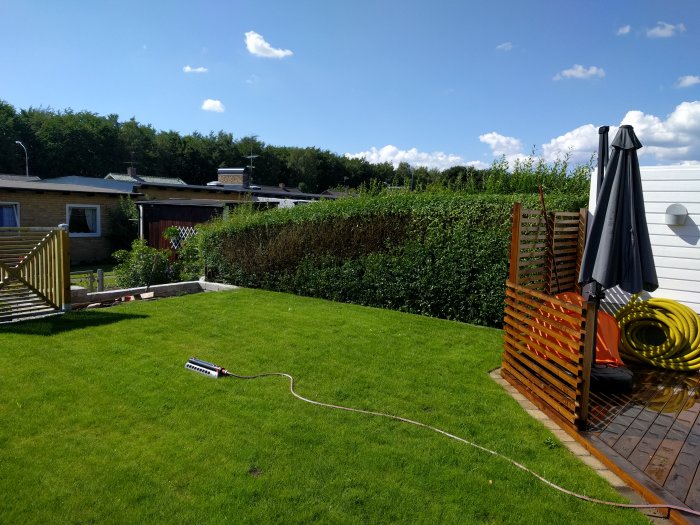 Lutande grön häck längs med trädgården, bredvid en terrass med parasoll och sopsäcksställning på gräsmattan.