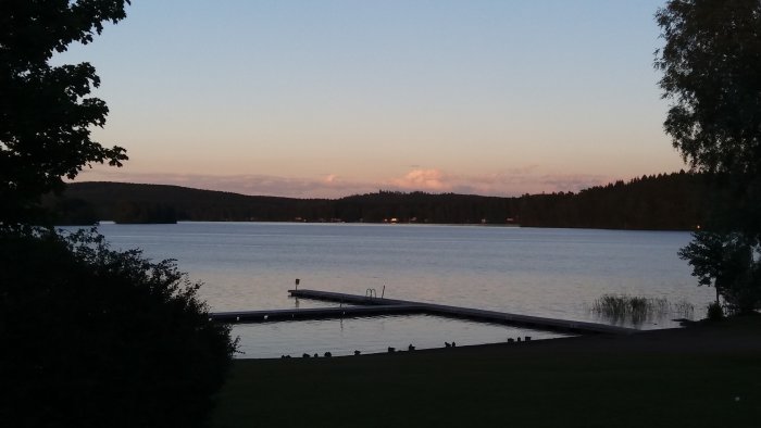 Kvällsutsikt över en sjö med brygga, träd och en skimrande vattenyta vid skymning.