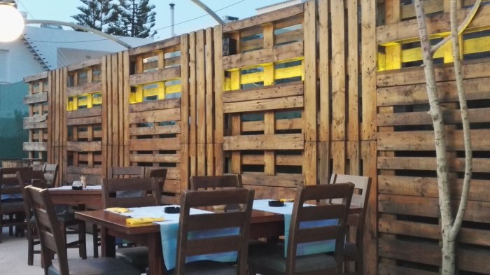 Återvunna lastpallar som använts för att skapa ett rustikt plankvägg, med gul belysning, vid en restaurangterrass.