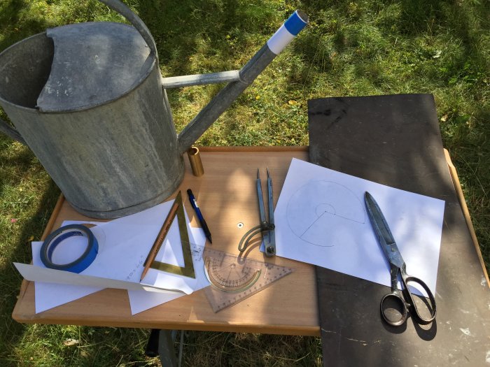 Verktyg och material för metallarbete inklusive mässingsbit, ritningar och vattenkanna på bord.