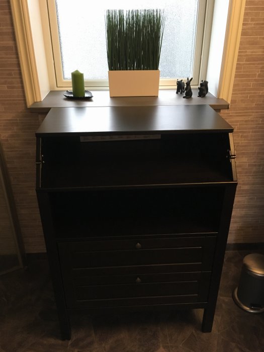 Ett monterat svart skötbord med stängd skötdyna framför ett fönster, dekorerat med en växt och ljus.