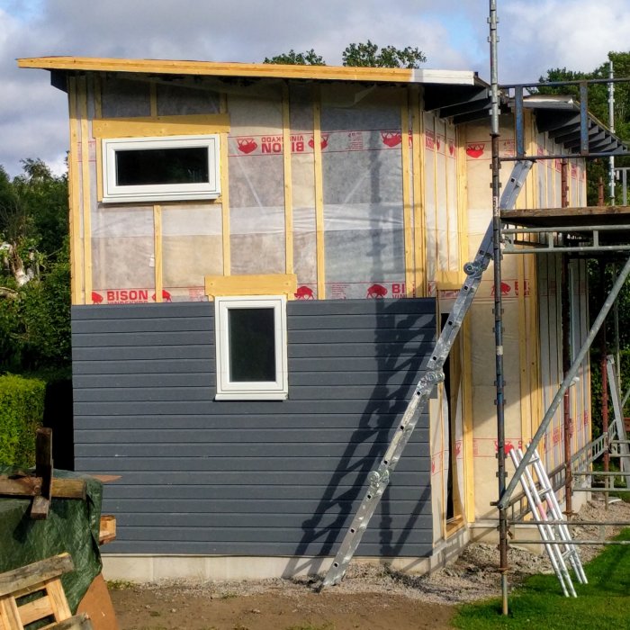 Delvis byggt hus med takstolar, regelverk, och några fönster på plats, samt delvis påsatt panel.