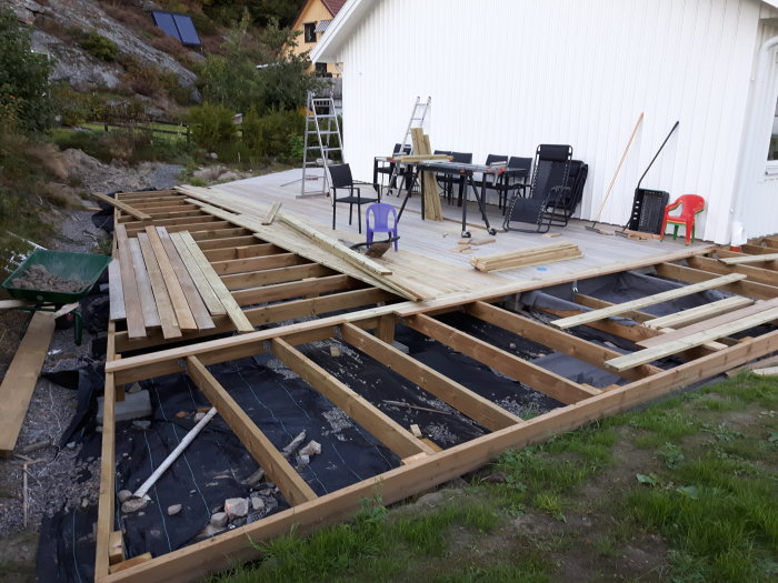 Pågående bygge av en stor altan vid hus, med träreglar, plankor och byggmaterial utspridda.