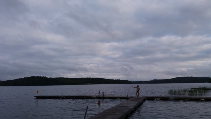 Molnig himmel över en sjö med personer på en brygga, en simmar och en annan står.