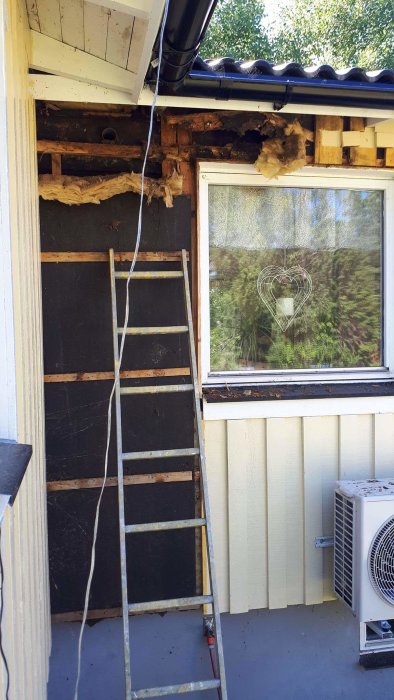 Husvägg med skador under takgräns där ett rör syns, stegar framför och luftvärmepump till höger.