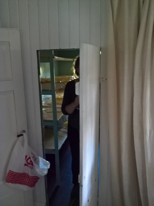 Person som öppnar dörr till rörigt rum med vit panel och trämaterial redo för förrådsrenovering.