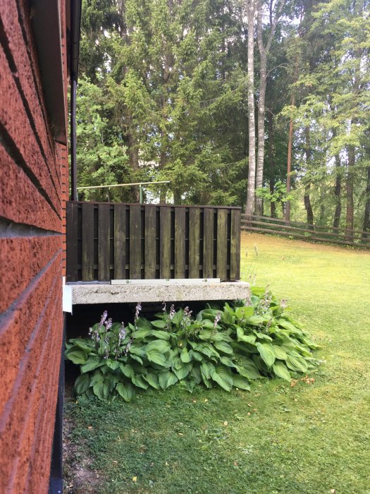 Trädgårdsbalkong med räcke och vattpass framför en tegelvägg, intill gröna buskar och träd i bakgrunden.