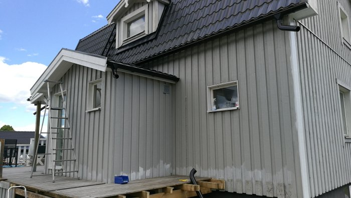 Ny målning på husvägg visar stark kontrast mot äldre blekt färg. Lång stege lutad mot väggen.