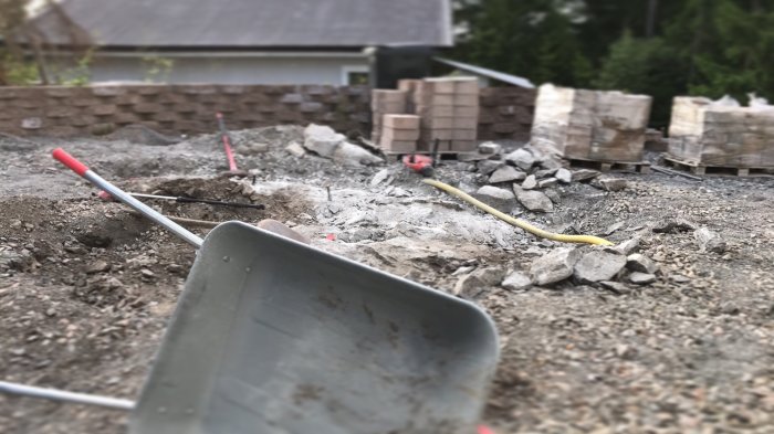 Bild på en uppbruten stenig mark med en skottkärra i förgrunden och staplar av markplattor i bakgrunden.