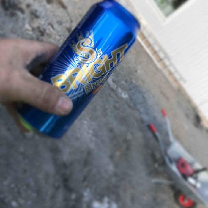En hand håller en ölburk i fokus med suddig bakgrund av en gräsklippare och trädgård.