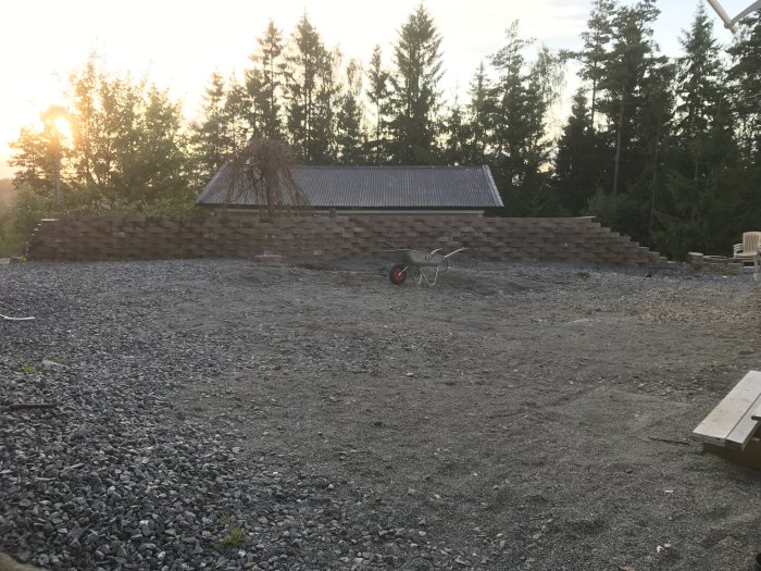 Förberedd grusyta för byggprojekt med stenmur och skottkärra i förgrunden, träd och solnedgång i bakgrunden.