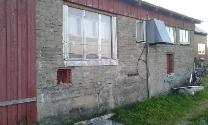 En gammal ladugård med slitna tegel och träväggar, sprickor och slitage syns tydligt, och en fråga om underhåll lämnas öppen.