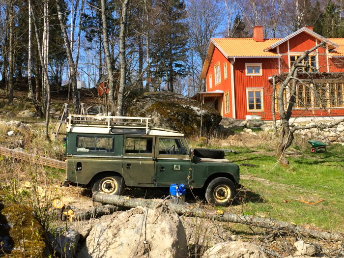 Nyrenoverat rött hus med vita knutar i soligt landskap, en klassisk grön Land Rover på förgrunden, stubbar och grönt gräs syns även.