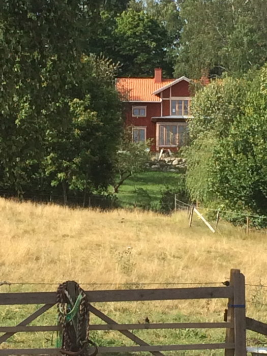 Nyfärdigmålat rött hus med tegeltak omgivet av gröna träd och ett gult gräsområde i förgrunden bakom ett trästaket.