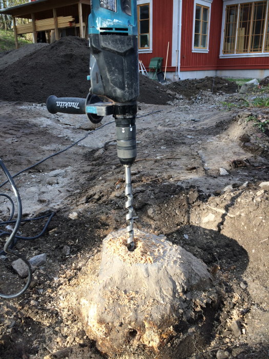 Borrmaskin används för att borra hål i trädstubbe under byggprojekt, med röd husfasad i bakgrunden.