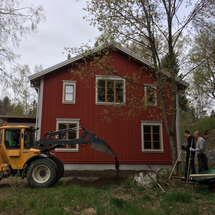 En färdigmålad röd timmerstuga under arbete med en grävmaskin och två personer som förbereder marken.