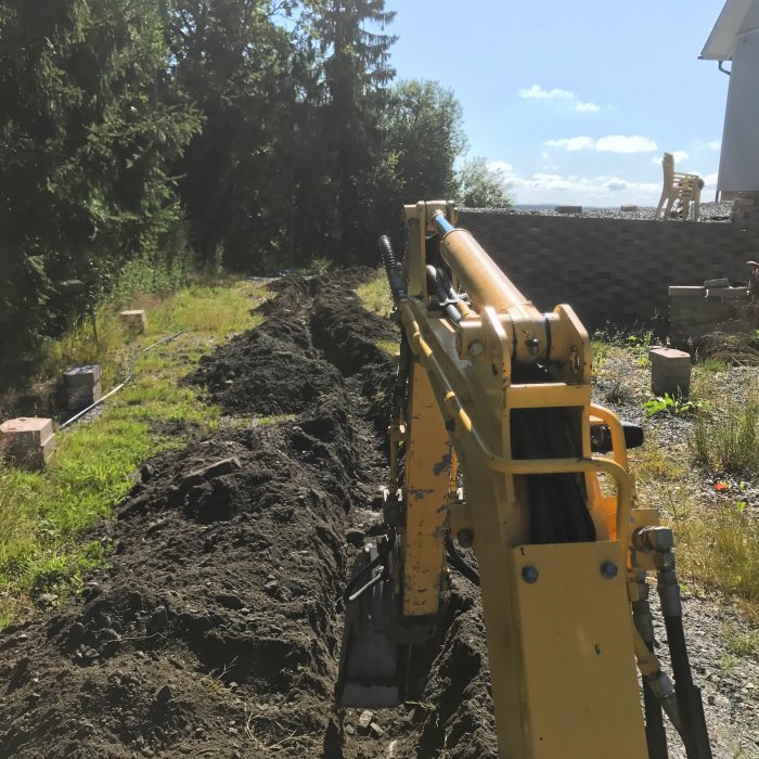 Gul grävmaskin gräver en lång dike i en trädgård för fiberkabelinstallation.