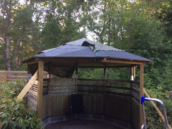 Pågående renovering av ett uterum med halvfärdigt tak och väggpanel av spårad trä, skyddad av svart plast, omgiven av grönska.