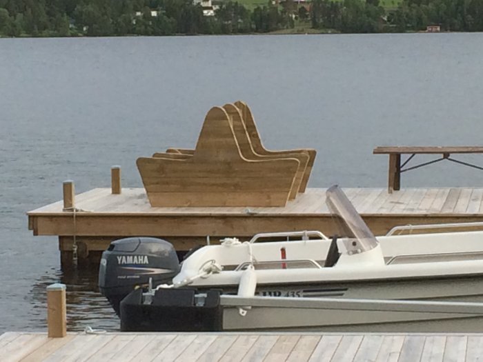 Obyggd träsoffa på en brygga vid en sjö med en båt och båtmotor i förgrunden.