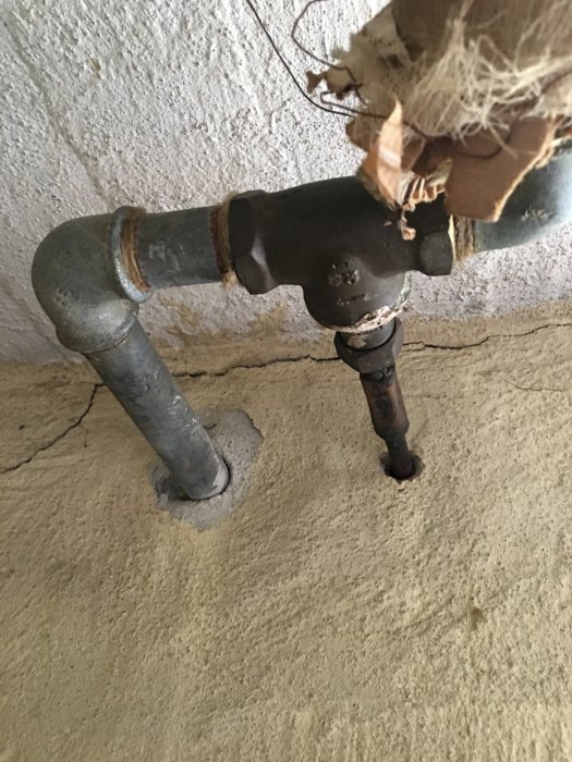 Äldre metallvattenrör och krök med isolering som går genom en betongvägg, en del av ett vattensystem i ett 60-talshus.