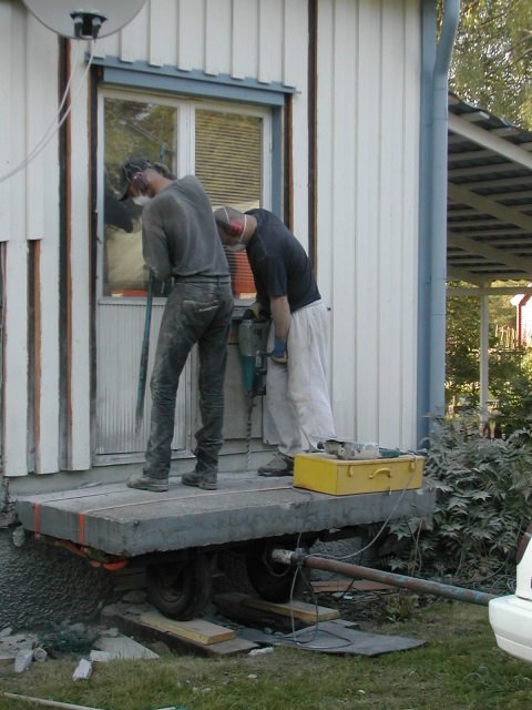 Två personer tar bort en betongplatta med en dollyvagn framför ett hus.