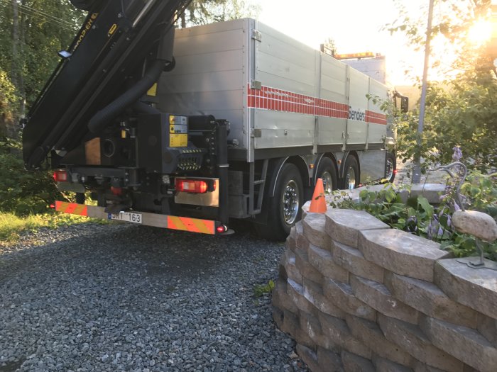 Lastbil med tippflak i en trädgård levererar grus tidigt, intill en stenmur.