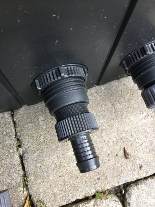 Svarta plastgängkopplingar som är fastskruvade på en yttre enhet liggande på ett stenunderlag.
