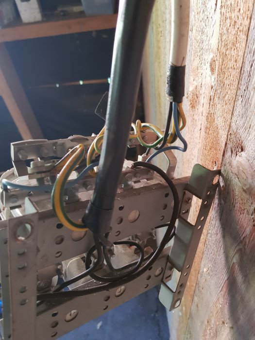 Äldre elinstallation med utsatta ledningar och öppen kopplingsdosa, fastsatt på en trävägg i ett uthus.