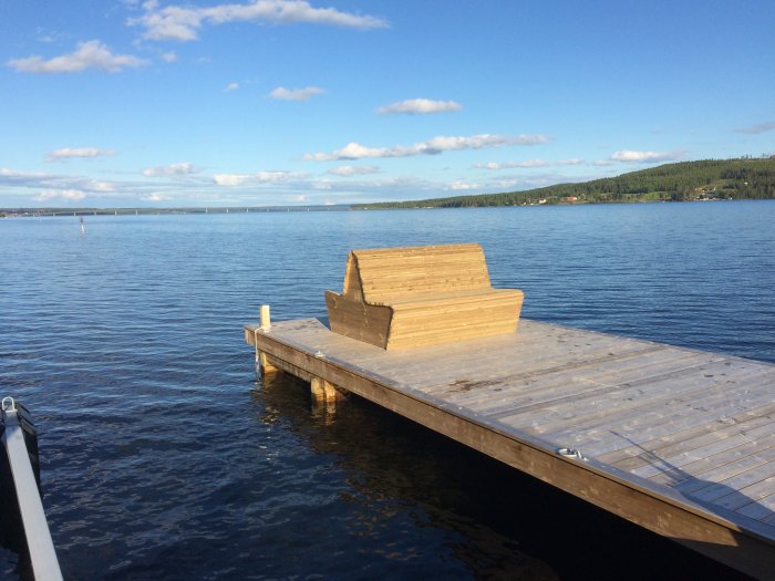 Nybyggd träbrygga vid sjö med oavslutad sittbänk under blå himmel.