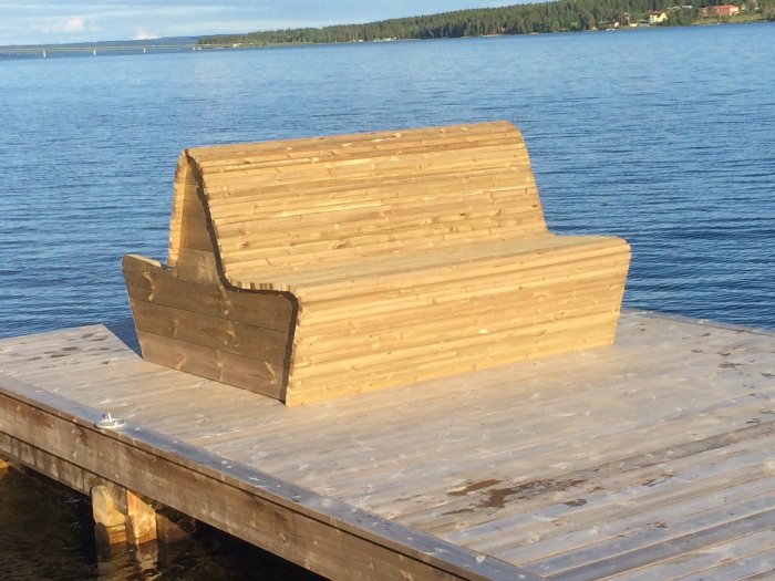 Träbänk under konstruktion på en brygga vid vattenkanten med en sjö i bakgrunden.