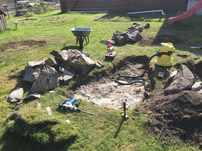 En trädgård med spräckta stenblock, grävmaskiner, en skottkärra och barnlekssaker på gräset.