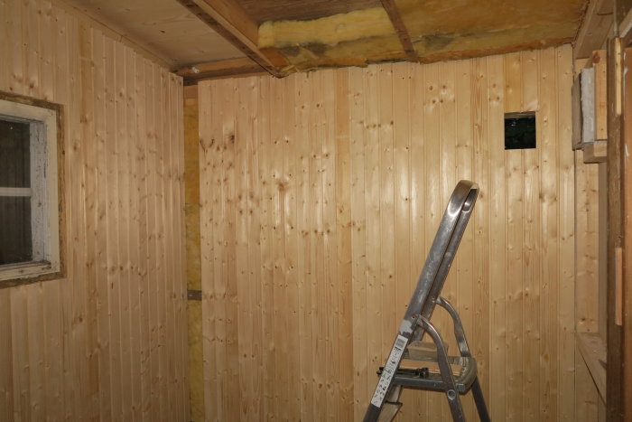 Nyuppsatt träpanel på väggar i ett byggrum med isolering synlig i taket och en stege, samt ett glasblocksfönster.