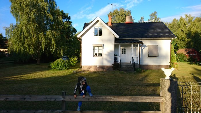 Ett hus med vit fasad och svart tak före renovering, med en person lutar mot ett staket i solnedgången.