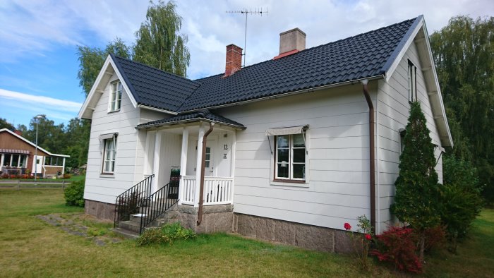 Vit villa med svart tak före renovering, omgiven av grönska och med nummer 12 på fasaden.